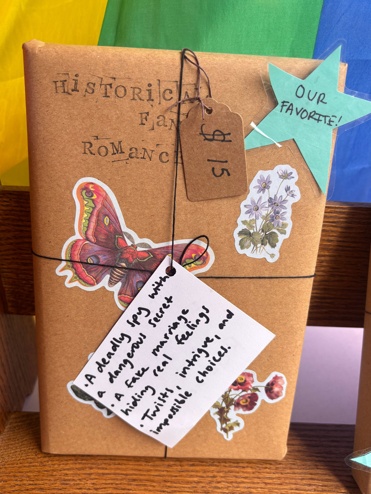 A wrapped book or journal in brown kraft paper with twine, decorated with stickers including a butterfly, flowers, and a handwritten note, displayed on a wooden shelf.