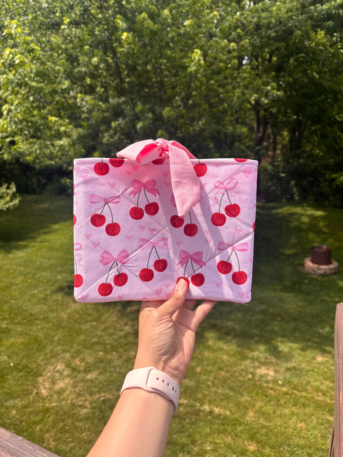 Hand holding a pink quilt with cherry pattern against a green outdoor background