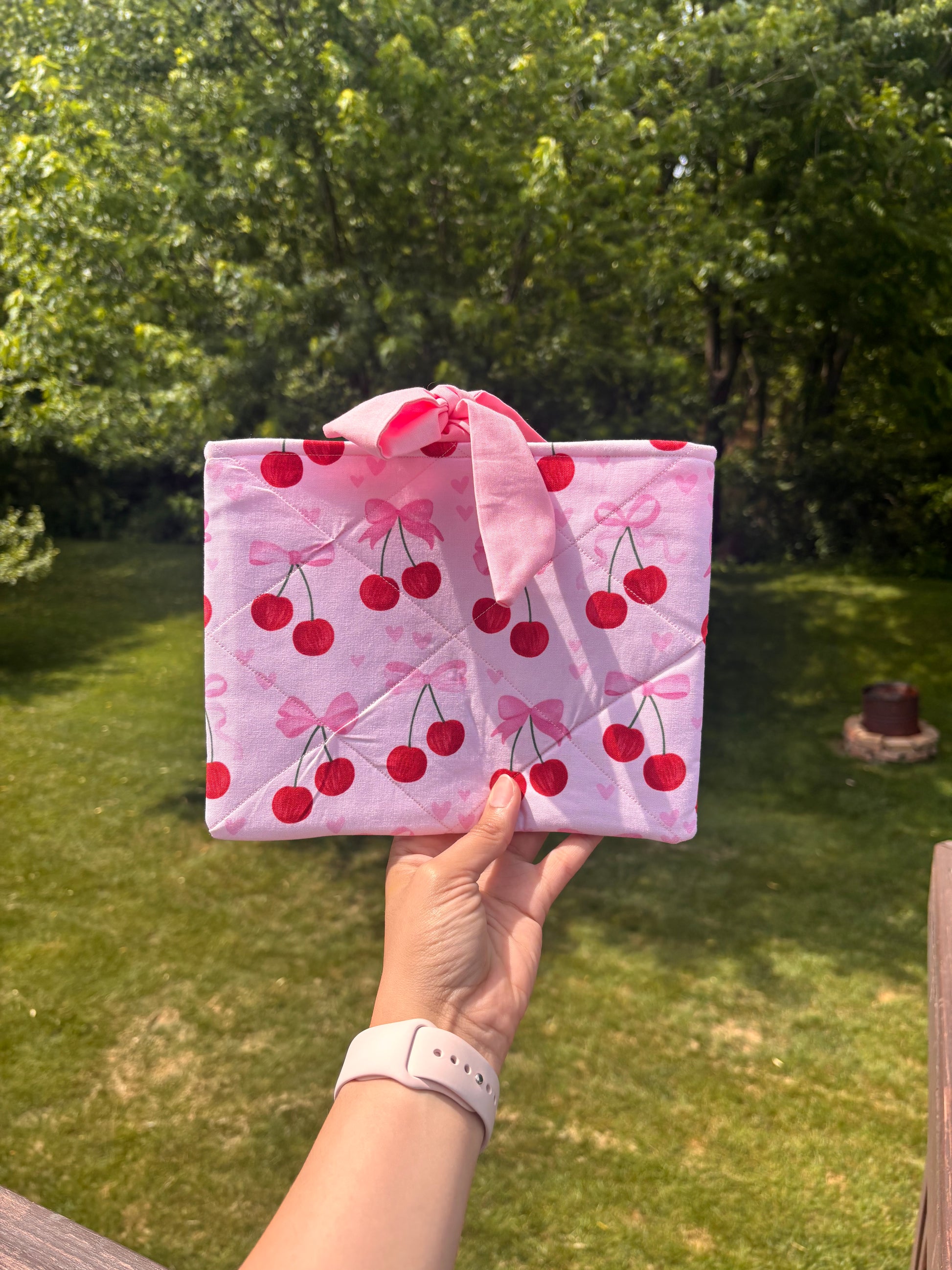 Hand holding a pink quilt with cherry pattern against a green outdoor background